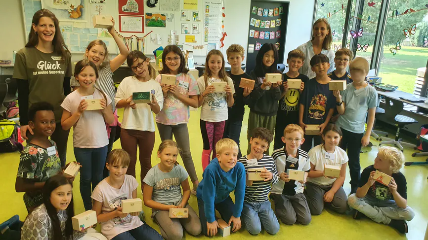 Langolino_Glücksworkshop_für_Kinder_Gruppenbild-16-9_866px_web Jessica Lang, eine Lehrerin und eine Gruppe von Grundschulkindern, die kleine Schatzkisten in den Händen halten
